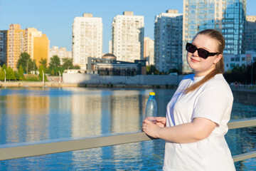 Portrait of a smiling young plus-size woman in sunglasses holding a plastic water bottle by a river with a modern cityscape behind her