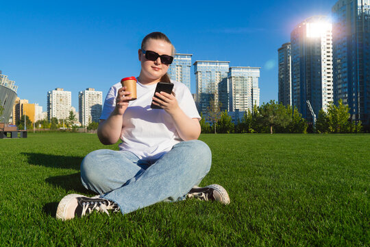 Young woman in sunglasses holding a phone and coffee cup while relaxing on green grass with modern skyscrapers in the background