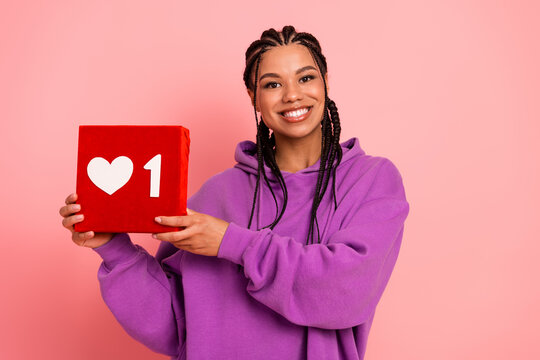 Cute woman in purple hoodie holding a red box with a heart and number symbol on a pink background