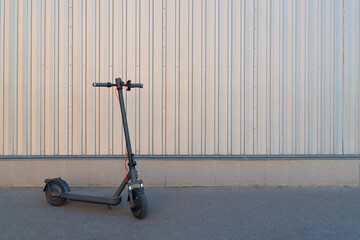 Contemporary black electric kick scooter standing alone on asphalt pavement next to a light corrugated metal fence on a sunny day