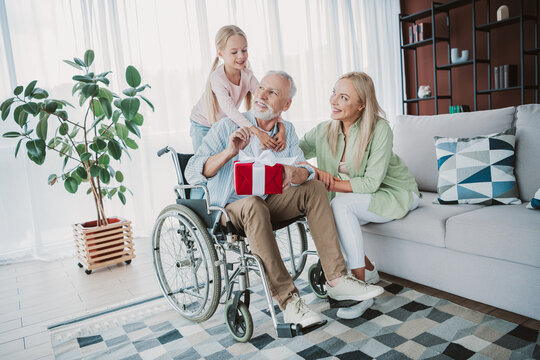 Fun loving family scene with grandfather in wheelchair receiving a gift from daughter and wife in a bright living room filled with plants cushions and a cozy home atmosphere - Powered by Adobe