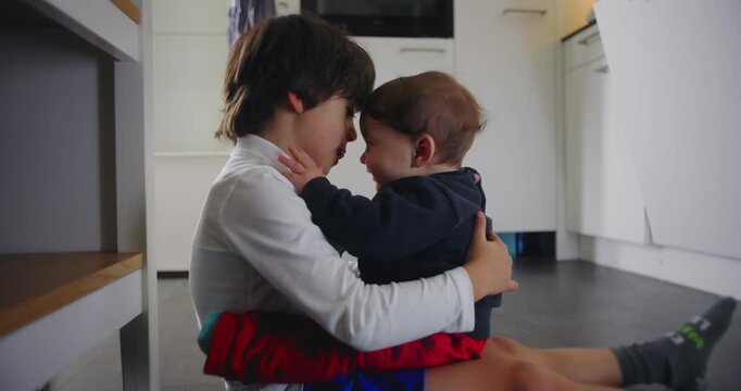 Older brother sitting on the kitchen floor hugging his baby sibling, both smiling and being playful, expressing tenderness, trust, deep emotional bond of family connection at home