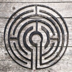 A rustic black labyrinth carved in wood, its mystical pattern standing out against aged gray planks &mdash; a symbol of journey, focus, and inner search.