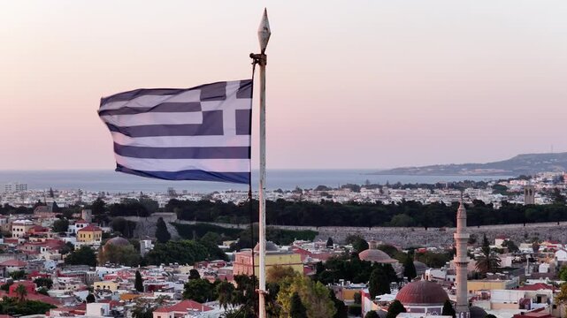 Greek flag waves above the city of Rhodes during a colorful dusk with views of the architecture and the distant sea