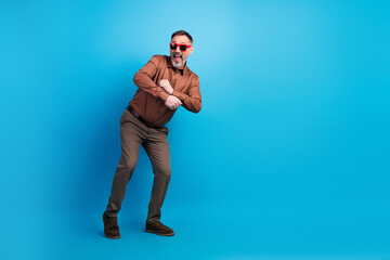 Energetic mature businessman dances playfully in a studio against a blue background wearing a brown shirt and red glasses