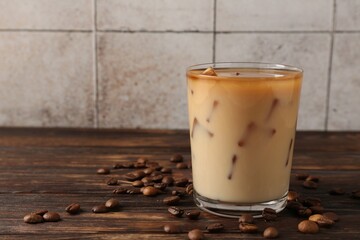 Tasty iced coffee with milk in glass and beans on wooden table against tiled wall, closeup. Space for text