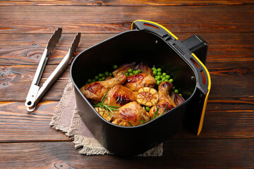 Air fryer basket with grilled chicken drumsticks, spices, green peas and tongs on wooden table, closeup