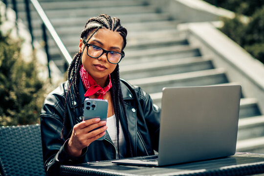 Young woman with braids using smartphone and laptop outdoors, enjoying her casual day in urban environment - Powered by Adobe