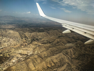 An airplane wing soars above a vast, arid mountainous landscape with scattered settlements under a...
