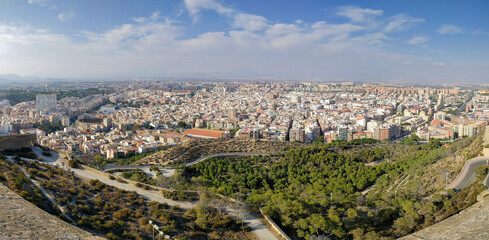 Obraz premium An expansive city panorama displays numerous buildings, winding roads, and green spaces under a partly cloudy sky.