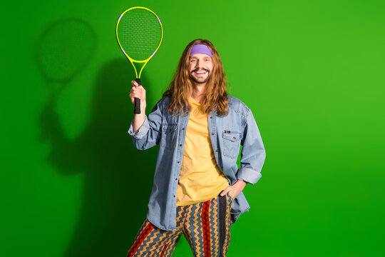 Young man with long hair in colorful retro outfit holding tennis racket and smiling against a vibrant green background