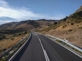 Spain - A winding asphalt road with white markings extends through a dry, mountainous landscape under a bright blue sky with scattered clouds.