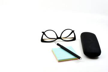Pen on a pad of reminder notes with a pair of women's glasses and a glasses case isolated on a plain white background. Copy space. No people.