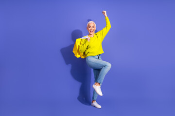 Fototapeta premium Cheerful mature woman in bright yellow sweater jumping with shopping bags against a purple backdrop ready for shopping and promotion