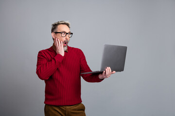 Surprised businessman holding a laptop against a gray background expressing amazement and curiosity...