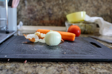 Peeled white onion with its skin, a whole carrot, and a tomato blurred in the background, sitting on a dark cutting board ready for chopping and cooking. Cooking. Recipes