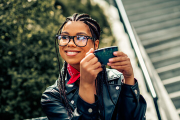 Young woman enjoying leisure outdoors with a cup of tea wearing braided hair, glasses, leather jacket, and scarf