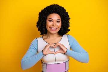 Smiling young woman with curly hair making heart shape gesture on yellow background portraying...