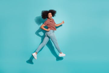 Young woman leaps in mid air against blue background wearing casual print shirt jeans and white...