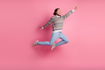 Cheerful young man jumping against a pink wall, wearing a colorful sweater and jeans, expressing...