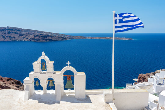 Greek orthodox church with bells and greek flag against famous white houses on Santorini island, Aegean sea, Greece.