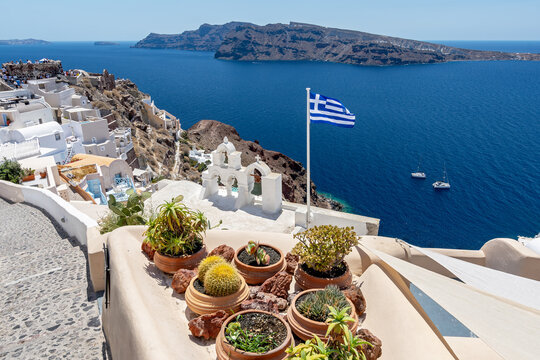 Greek orthodox church with bells and greek flag against famous white houses on Santorini island, Aegean sea, Greece.