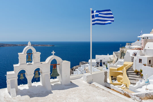 Greek orthodox church with bells and greek flag against famous white houses on Santorini island, Aegean sea, Greece.