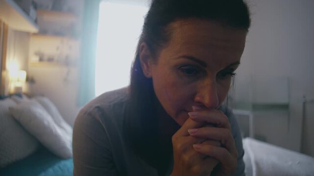 Woman sitting alone on bed, hands clasped and head lowered, expressing emotional exhaustion, quiet reflection, and sorrow in soft natural light home setting