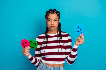 Young woman with braids holds colorful emotion faces against blue background wearing striped top and denim jeans