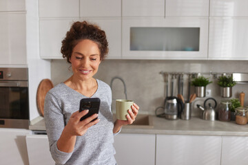 A woman stands in her modern kitchen, smiling as she checks her smartphone with a cup of coffee in her other hand, showcasing a relaxed morning routine. © Stockphotodirectors