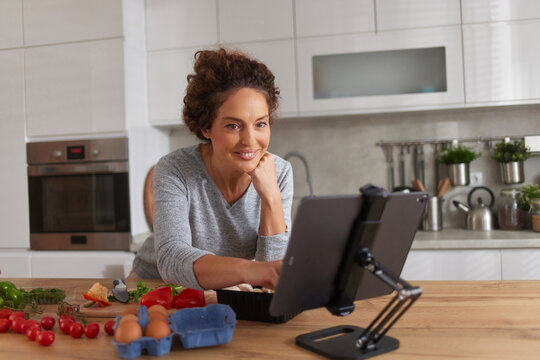 A woman smiles as she prepares a meal in her bright kitchen. Fresh ingredients like tomatoes and eggs surround her while she follows a recipe on a tablet. - Powered by Adobe