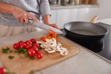 A person is chopping colorful bell peppers, cherry tomatoes, and mushrooms on a wooden cutting board in a bright kitchen. A frying pan sits on the stove, ready for cooking.