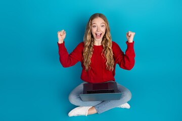 Adorable young woman in a red sweater celebrating success with laptop on vibrant blue background