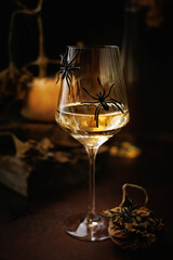 White wine glass with black spiders on table near dried leaves and pumpkin in Halloween still life