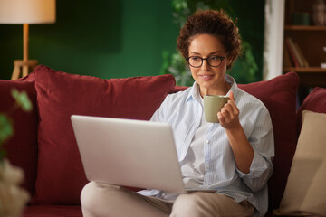 A relaxed woman sits on a cozy couch, sipping a warm drink from a green cup while focused on her...