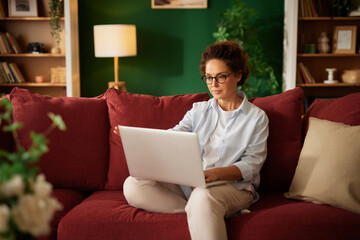 A woman sits comfortably on a red couch, focusing on her laptop while surrounded by plants and warm...