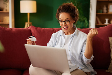 Woman with curly hair and glasses happily shops online with her credit card while seated on a red...