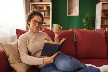 A woman relaxes on a red sofa, reading a book while holding a mug. She wears a soft sweater and...