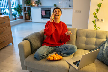 Joyful laughing woman chats on phone while peeling mandarins and using laptop on sofa. Happy mature female multitasking, citrus snack break, relaxed home office, laughing senior during remote work