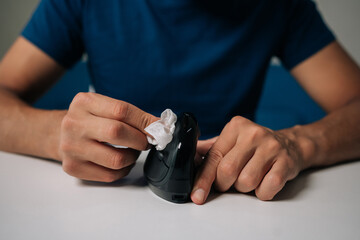 Cropped shot of unrecognizable man cleaning modern black vertical mouse with disinfectant wipe, promoting hygiene and preventing spread of germs, sitting at white table in workplace or home office.