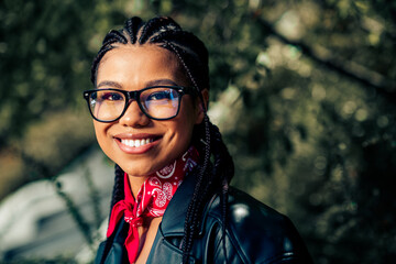 Confident young woman with braided hairstyle and glasses outdoors, adorned with a vibrant red scarf and leather jacket