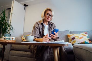 Middle aged woman reviewing expenses, managing personal finances on smartphone mobile app while sitting on couch with coffee in cozy living space. finance planning, financial balance check, budgeting