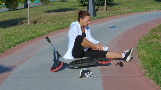 Pain from a scooter accident. Woman sitting on the ground, gripping her ankle in pain after a scooter accident in a park. The scene captures the aftermath of a fall, highlighting injury and distress