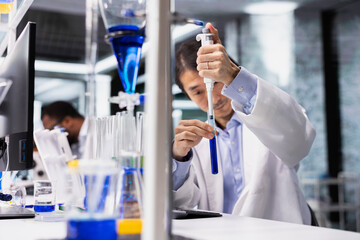 Senior analytical chemist uses pipette to transfer liquid into test tube containing blue solution at laboratory bench. Asian man in research facility does pipetting procedure with chemical reagent
