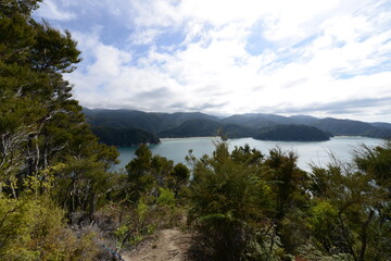 Obraz premium Panoramic view from hiking trail overlooking a protected bay with turquoise waters, forested islands, and distant mountains under a partly cloudy sky, showcasing pristine coastal wilderness.