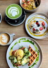 Stylish brunch with waffles, eggs benedict, pancakes, matcha latte, and cappuccino on a wooden table.