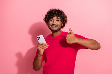 Smiling young man in red t-shirt showing thumbs up gesture with modern smartphone on pink background