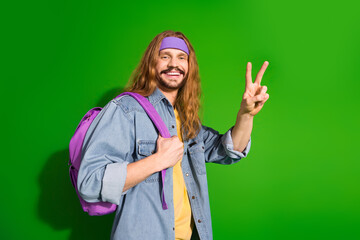 Smiling young man with long hair showing a peace sign gesture while wearing denim over a green...