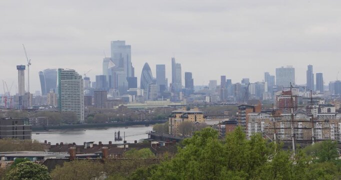 Wide elevated view of London under an overcast sky. The River Thames curves past rooftops, cranes and financial towers, showing expansion, housing pressure and unequal growth at the edges of the city.