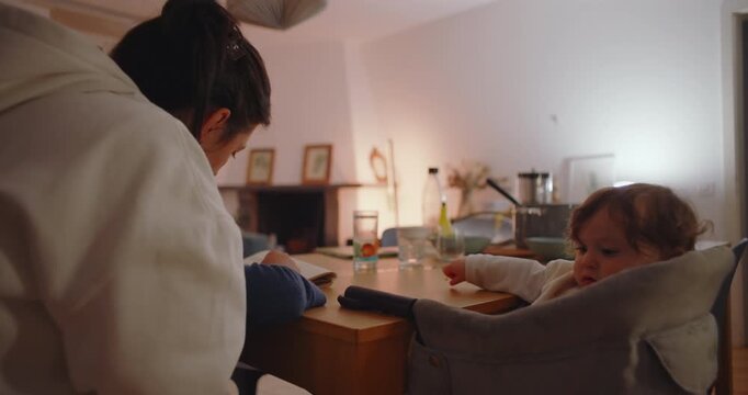 Mother leaning over dining table guiding son with schoolwork while younger sibling sits in high chair holding red toy, symbolizing multitasking parenting at home